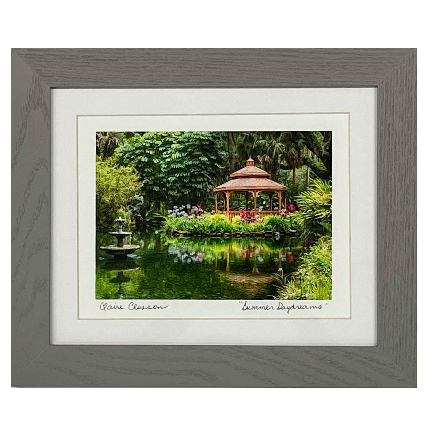 ECC “Summer Daydreams” Framed Print by Photographer Claire Closson. Stunning photograph of a gazebo on the water surrounded by flowers and lush green. A tiered fountain cascades water. Soft reflections in the water. A 5 x 7 print framed to 8 x 10 inches under glass. Matted in white with a gray wooden frame.