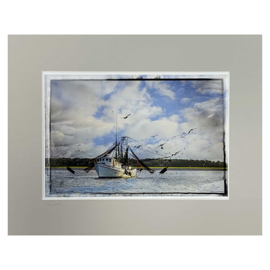 Photograph of a shrimp boat with seagulls and pelicans flying around it.  The sky is cloudy and the boat has land behind it.  In a white mat.