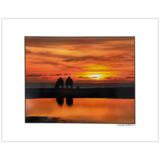 Two silhouetted people sitting in chairs on the water at Treasure Island with an orange sunset sky and reflection in the water. Shown in a white mat with a black core. Signed by the artist.