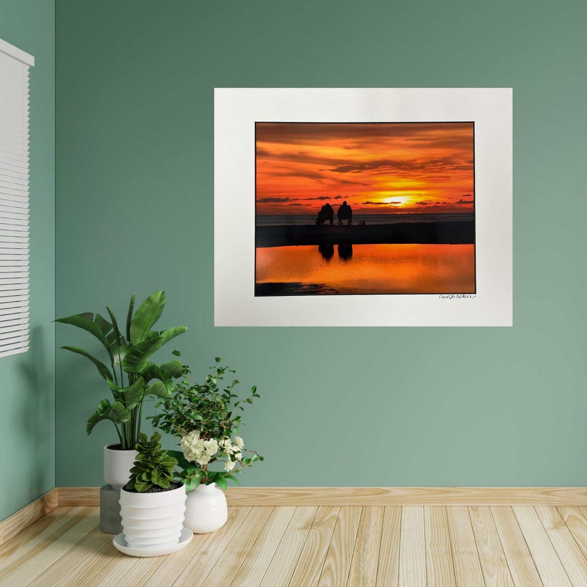 Two silhouetted people sitting in chairs on the water at Treasure Island with an orange sunset sky and reflection in the water. Shown in a white mat with a black core. Signed by the artist. Hanging in a room with a green wall and wood floor.