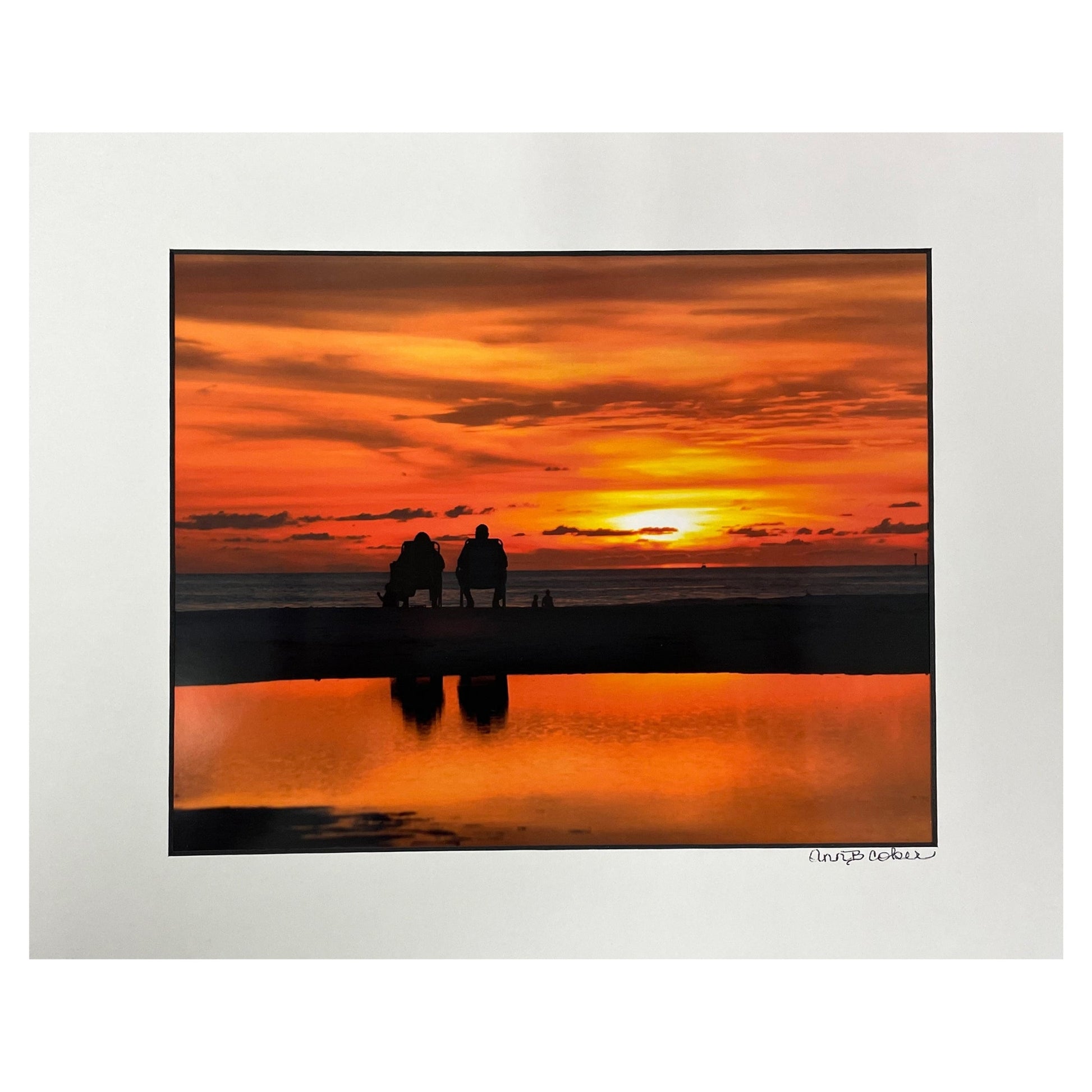 Two silhouetted people sitting in chairs on the water at Treasure Island with an  orange sunset sky and reflection in the water.  Shown in a white mat with a black core.  Signed by the artist.